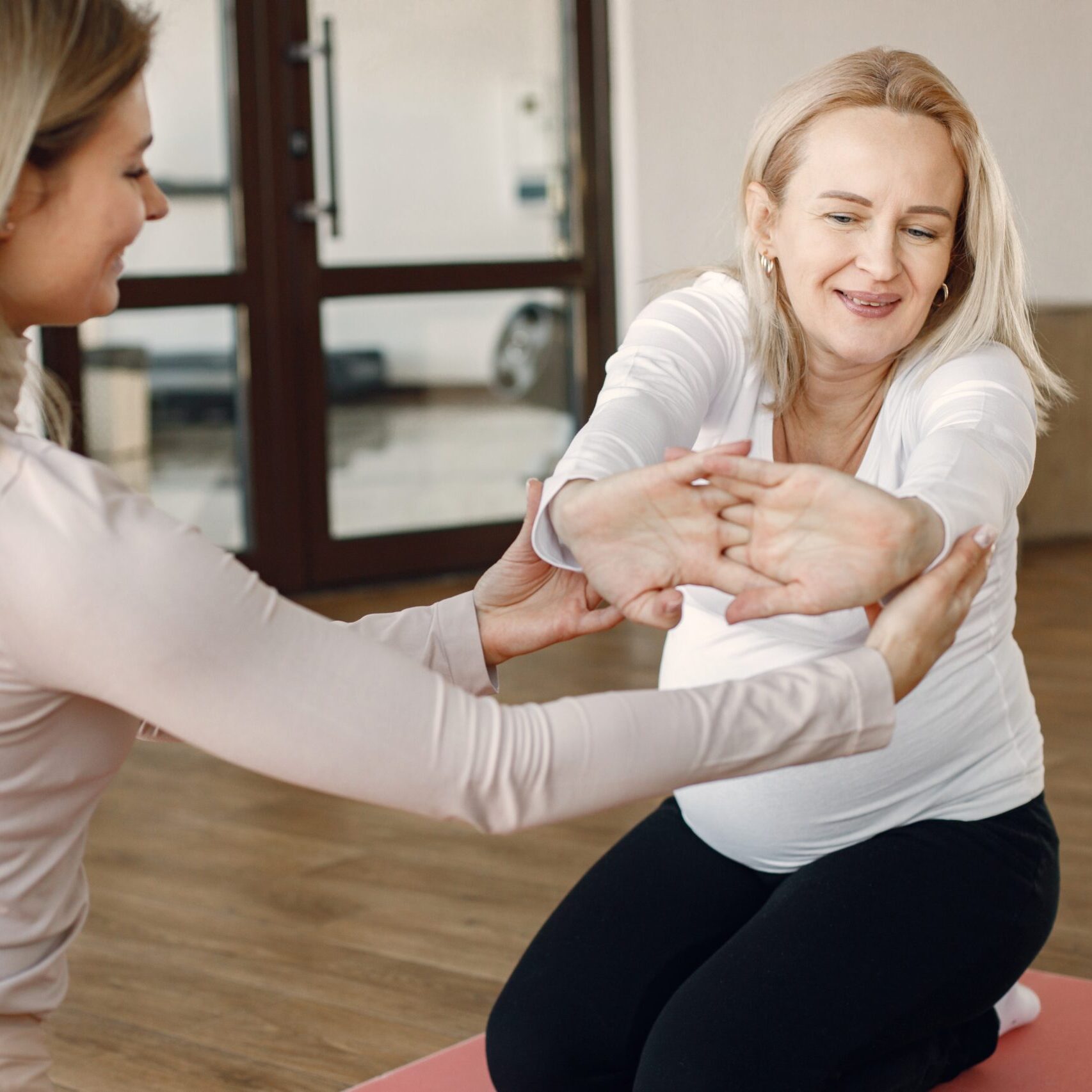 Pregnant woman doing yoga with personal trainer. Yoga trainer assisting pregnant woman while doing exercises. Blonde pregnant woman wearing white clothes.